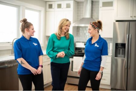 Two Jeannie Cleaning team members in blue uniforms chat and smile with a client in a bright, clean kitchen.