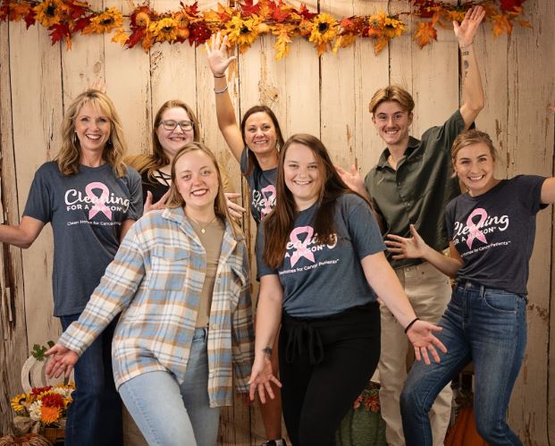 A cheerful group photo of a cleaning team standing in front of a rustic wooden backdrop decorated with vibrant autumn leaves. The team members are smiling and posing energetically, wearing casual outfits and "Cleaning for a Reason" shirts, showing camaraderie and enthusiasm.